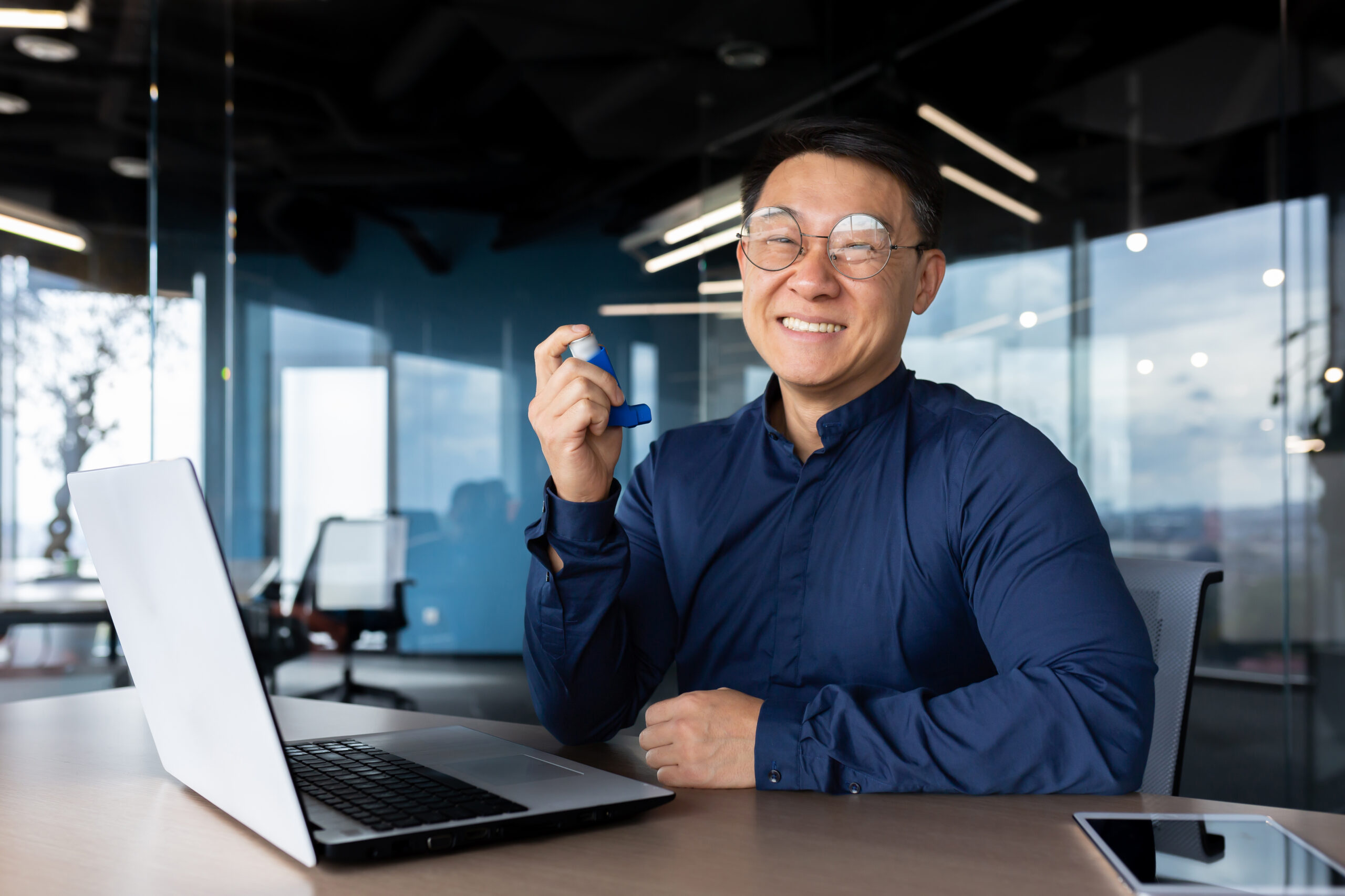 A young Asian businessman, freelancer, programmer works in the office at a laptop. He holds an asthma and allergy inhaler in his hands, smiles at the camera, poses, advertises, recommends.